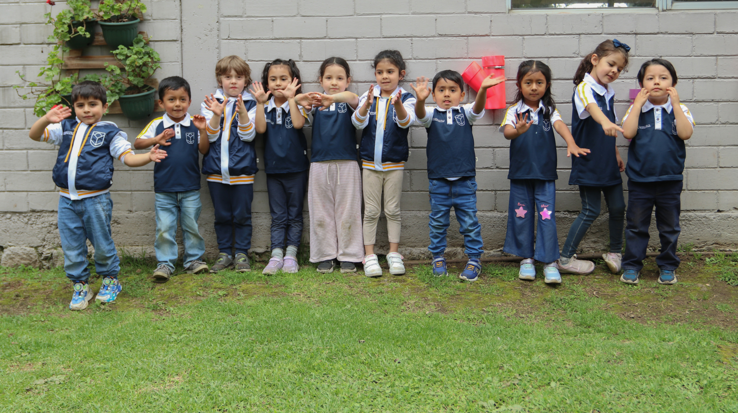 Niños y niñas pequeños de Comuna con uniforme saludando frente a una pared en el jardín